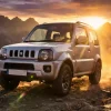 ゴールデンアワーの夕暮れ時に、荒れた未舗装路を走行するシルバーのジムニーJB23を撮影した劇的な画像です。車体には使い込まれた様子を示す泥の跡があり、タフで頼れる印象を与えています。背景には雄大な山脈のシルエットが広がり、シネマティックなサンフレア効果と黄金色の光線が、フロントグリルとヘッドライトを美しく照らし出しています。